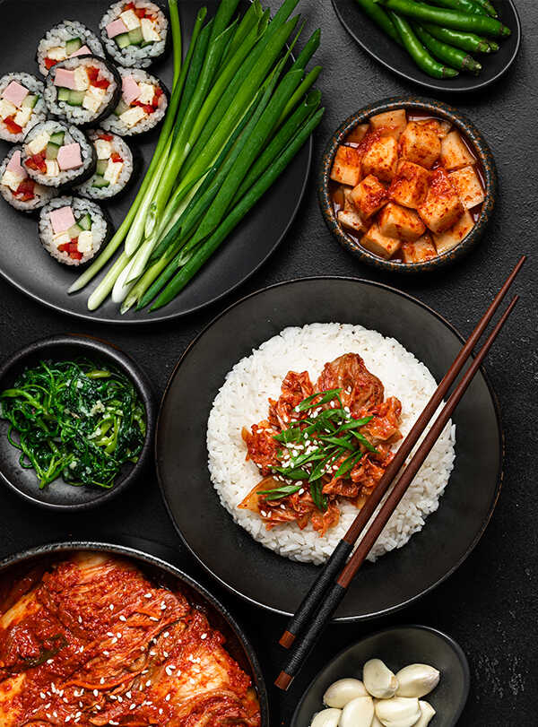 Una variedad de platos coreanos sobre una superficie oscura: un bol de arroz cubierto con kimchi y cebollas verdes, rollitos de kimbap, cebolletas, kimchi, judías verdes, espinacas y dientes de ajo. Los platos están dispuestos en cuencos y platos negros.
