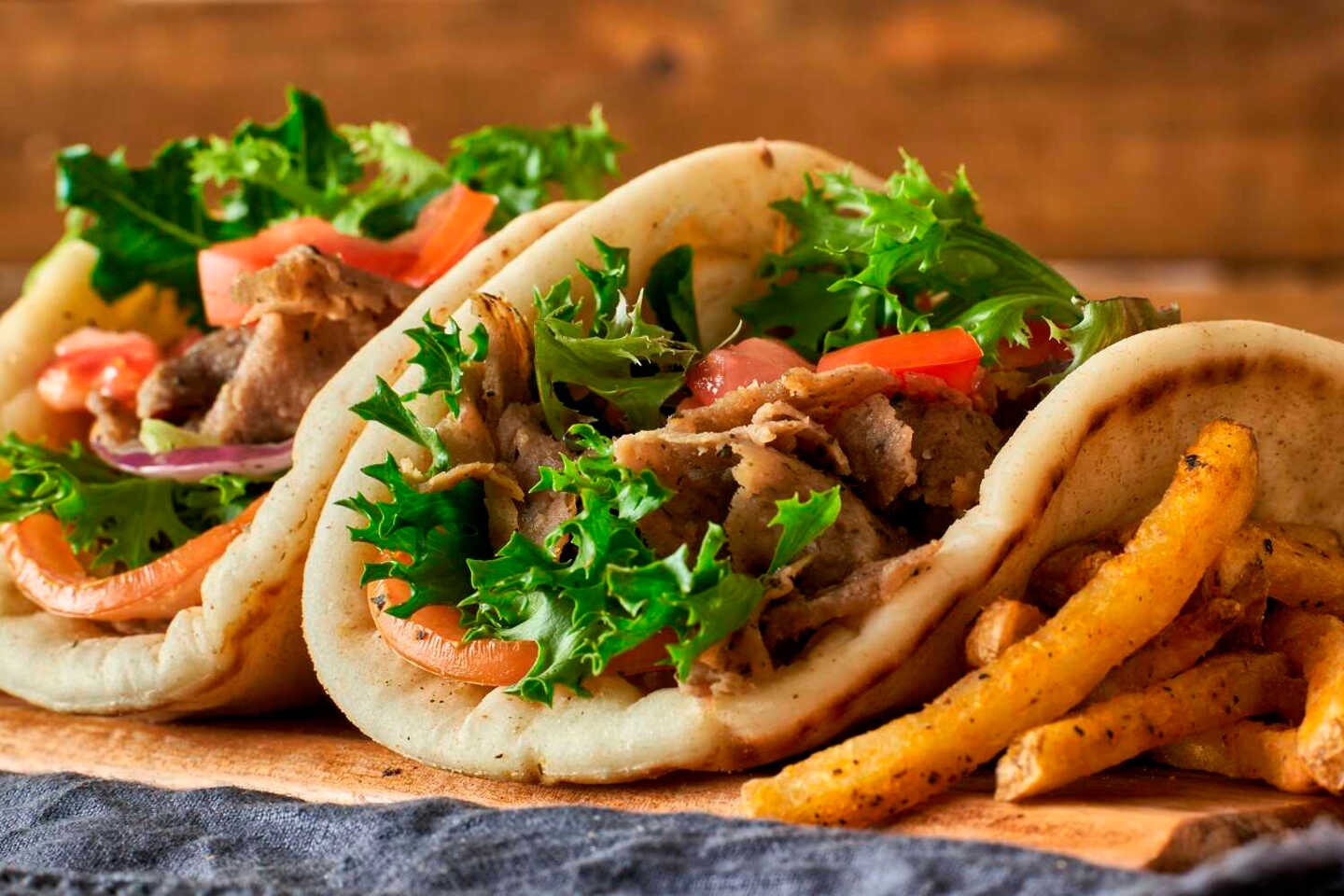 Dos wraps de pita rellenos de carne cortada en rodajas, lechuga y tomate, colocados sobre una tabla de madera con una ración de patatas fritas sazonadas. El fondo tiene un aspecto rústico, desenfocado y de tonos cálidos. Dos wraps de pita rellenos de carne cortada en rodajas, lechuga y tomate, colocados sobre una tabla de madera con una ración de patatas fritas sazonadas. El fondo tiene un aspecto rústico, desenfocado y de tonos cálidos.