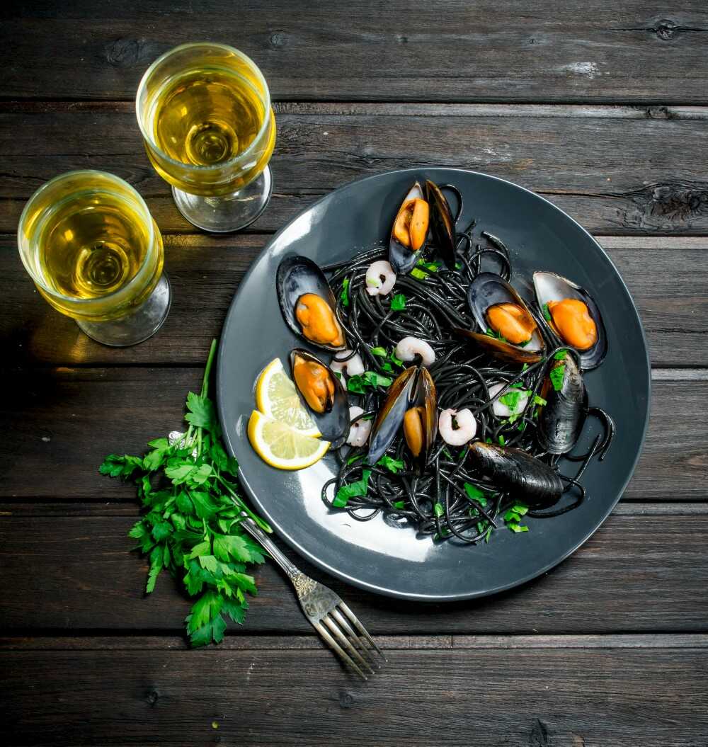 Un plato de pasta con mariscos, fideos negros, mejillones y camarones sobre una mesa de madera oscura. Decorado con perejil y rodajas de limón. Junto al plato hay dos copas de vino blanco, con un tenedor y una ramita de perejil cerca.