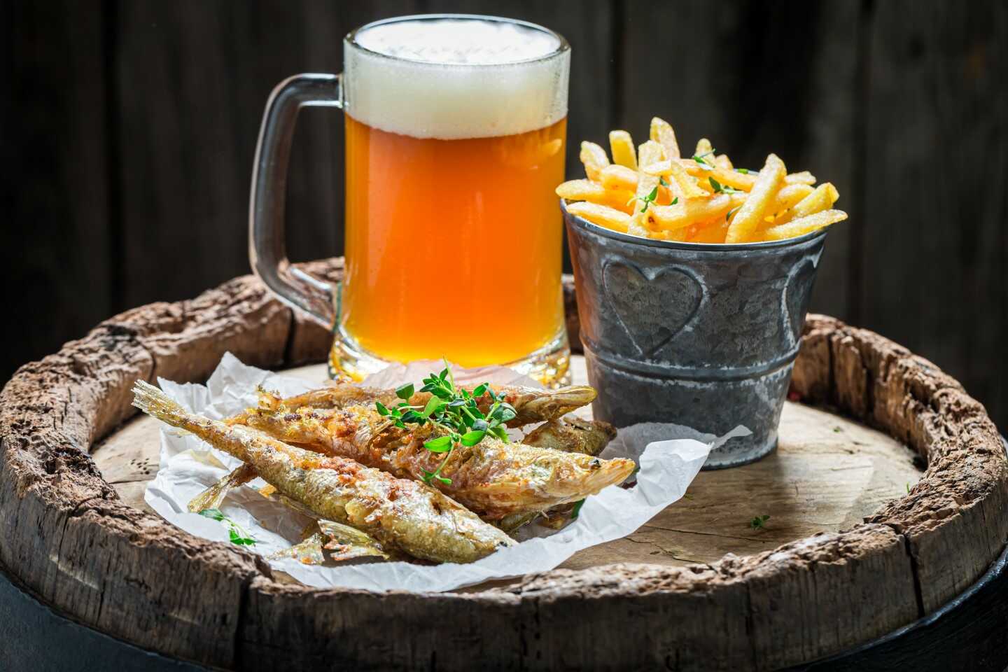 Una jarra de cerveza espumosa se encuentra junto a unas patatas fritas crujientes en una taza de metal rústica y una ración de pescado pequeño a la parrilla sobre papel de pergamino, todo ello colocado en un barril de madera. Adornado con hierbas, el ambiente es acogedor y rústico. Una jarra de cerveza espumosa se encuentra junto a unas patatas fritas crujientes en una taza de metal rústica y una ración de pescado pequeño a la parrilla sobre papel de pergamino, todo ello colocado en un barril de madera. Adornado con hierbas, el ambiente es acogedor y rústico.