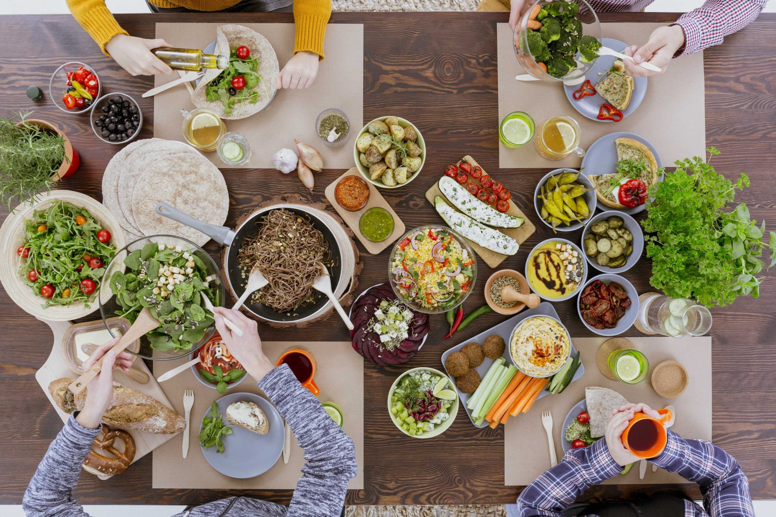 Vista superior de una mesa de madera llena de platos vegetarianos coloridos, que incluyen ensaladas, sándwiches, verduras a la parrilla, encurtidos y hierbas. Cuatro personas están sentadas alrededor de la mesa, preparando y compartiendo la comida.