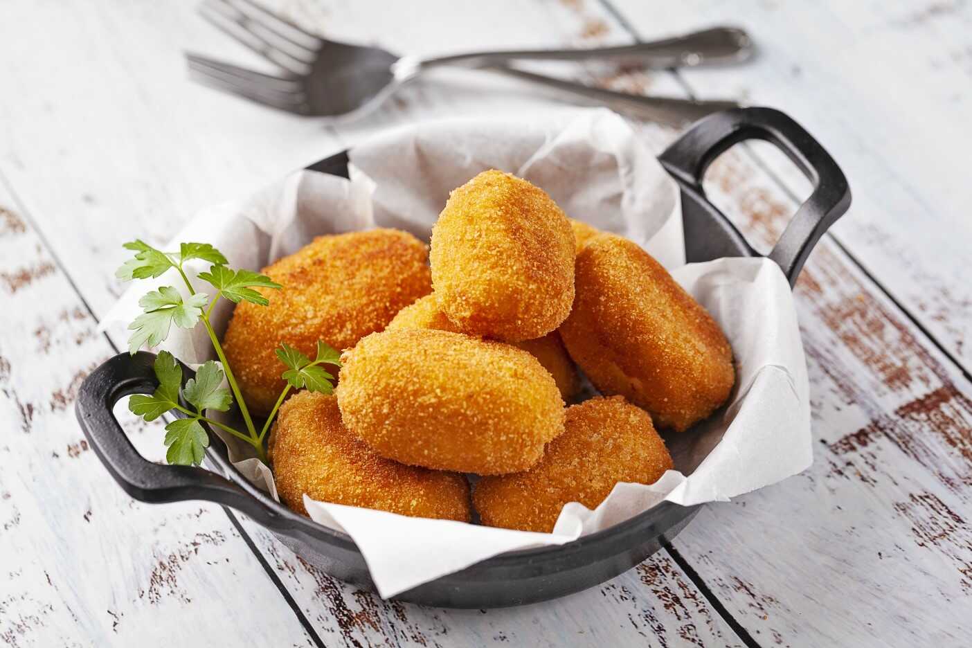 Una sartén negra con croquetas doradas sobre papel de pergamino blanco, adornadas con perejil. Tres tenedores yacen en el fondo sobre una superficie rústica de madera blanca. Una sartén negra con croquetas doradas sobre papel de pergamino blanco, adornadas con perejil. Tres tenedores yacen en el fondo sobre una superficie rústica de madera blanca.