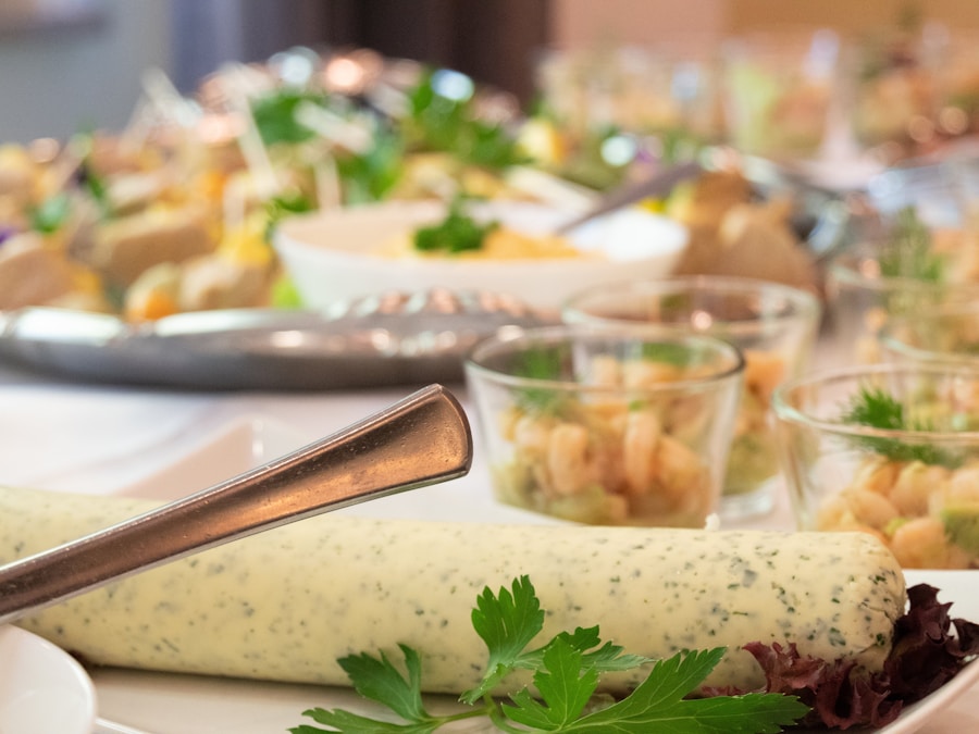 Primer plano de una mesa de buffet con un trozo de mantequilla con hierbas en primer plano. Al fondo, hay pequeñas copas de cristal con aperitivos, adornadas con hierbas y rodeadas de varios platos de comida.