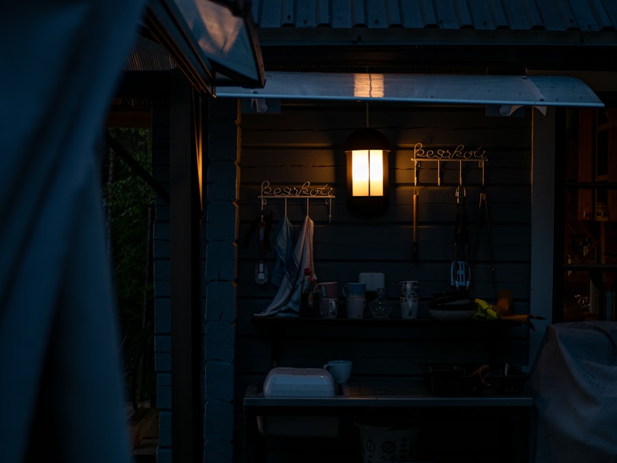 Una cocina al aire libre con poca luz al anochecer, con una luz de pared brillante sobre un estante con utensilios y toallas colgados. Varios artículos de cocina están dispersos sobre una encimera debajo de un toldo de metal.