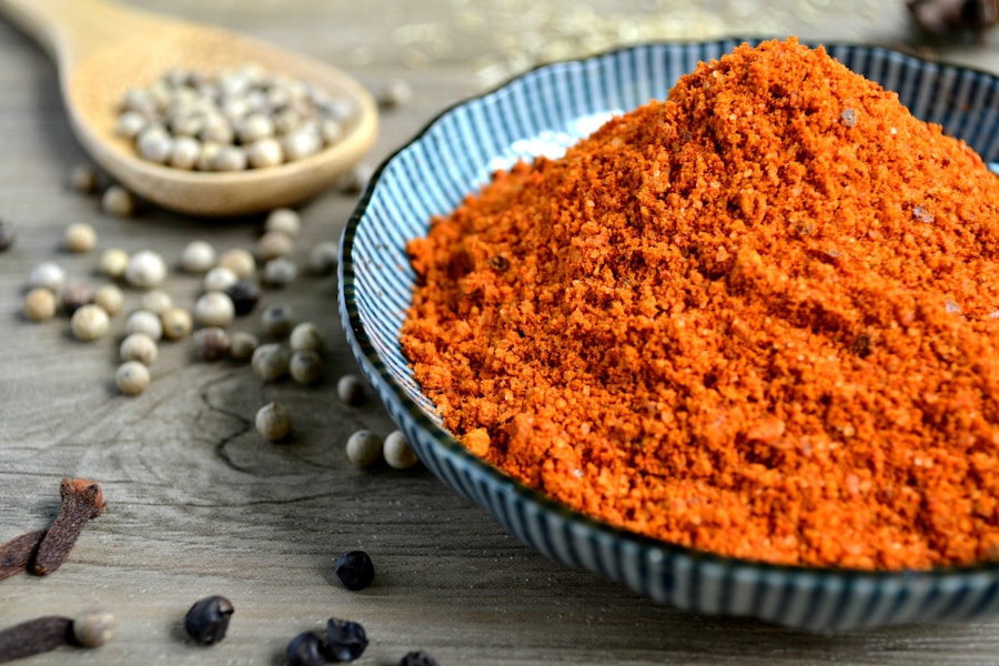 Sobre una superficie de madera se coloca un cuenco azul lleno de chile en polvo de color rojo anaranjado intenso. Alrededor del cuenco hay granos de pimienta blanca esparcidos y una cuchara de madera llena de más granos de pimienta, lo que crea una escena rústica y llena de sabor.