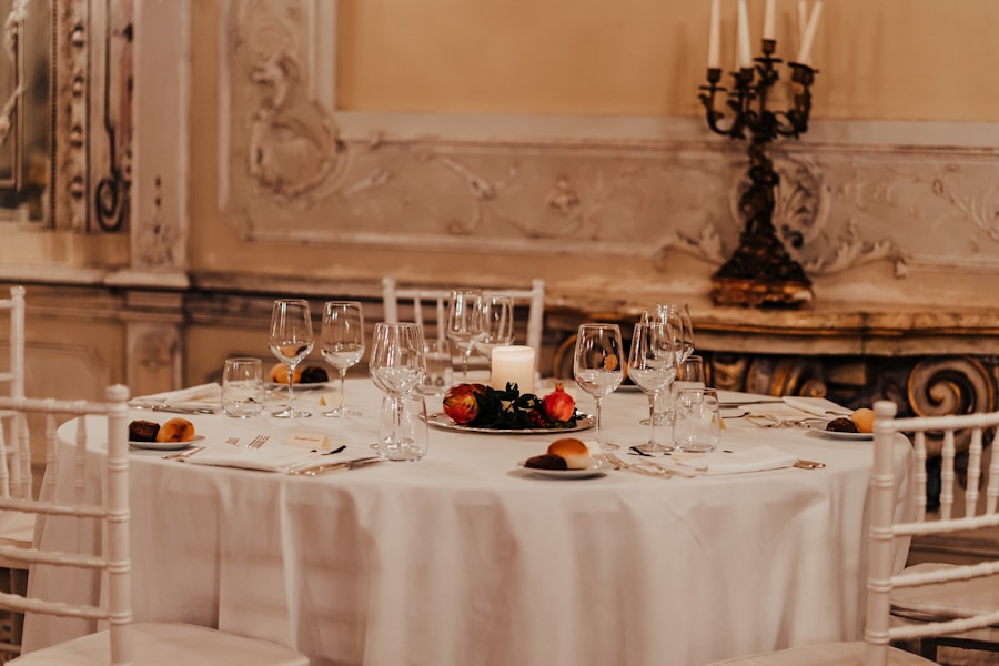 Una mesa redonda preparada para una cena formal con un mantel blanco, rodeada de sillas blancas. Tiene copas de vino, velas, un centro de mesa con frutas y panecillos. La pared ornamentada y los candelabros añaden elegancia al ambiente.