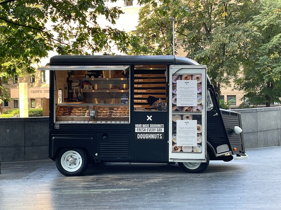 Un food truck vintage negro estacionado junto a una pasarela, exhibiendo una variedad de donas tras un cristal. Un letrero lateral dice: "Donas artesanales frescas todos los días". Se ven árboles y un edificio al fondo.