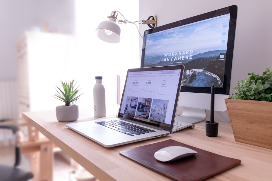 Un escritorio ordenado con una computadora portátil, un monitor de computadora que muestra "TRABAJA DURO EN CUALQUIER LUGAR", un mouse inalámbrico sobre un mouse pad, una botella de agua, una pequeña planta en maceta, una tableta gráfica y una planta decorativa, en una habitación luminosa.