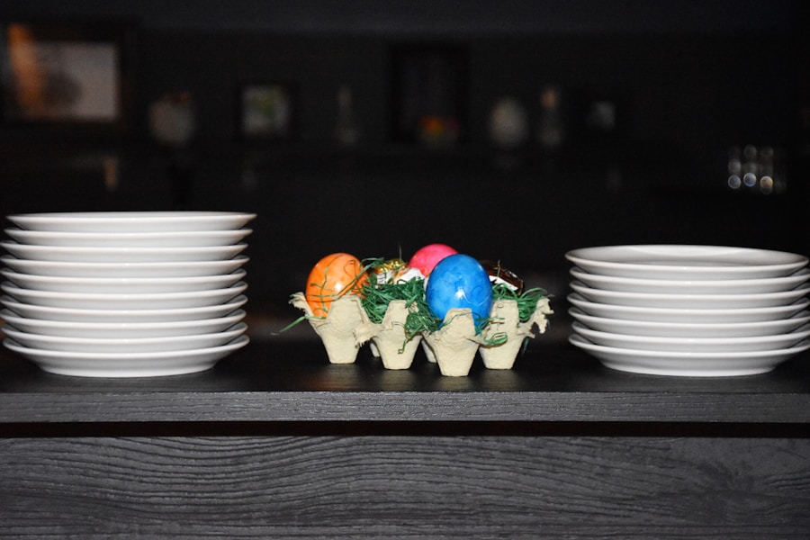 A cada lado de una caja de cartón hay dos pilas de platos blancos que contienen huevos de Pascua coloridos y césped verde, todo colocado sobre una superficie oscura con un fondo borroso.
