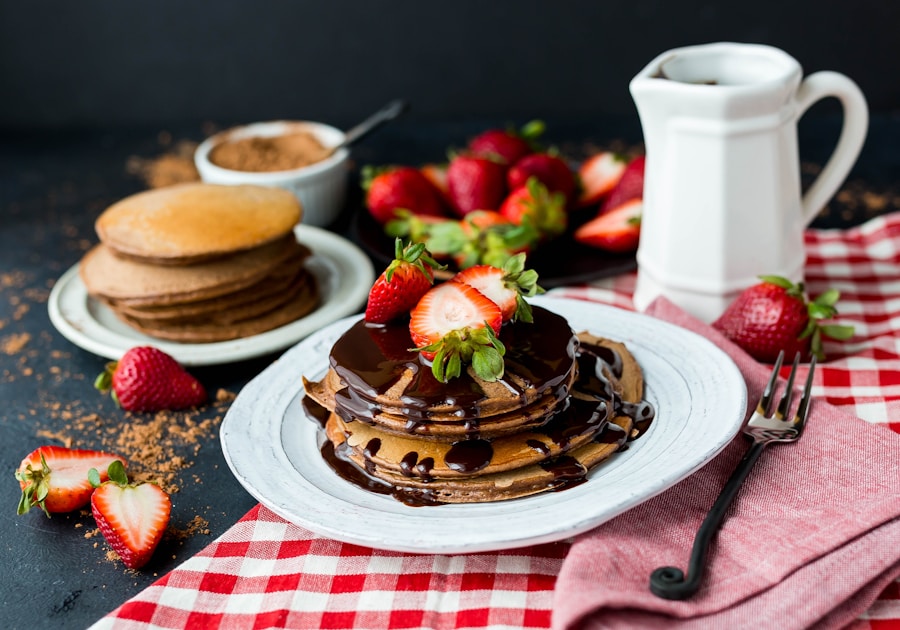 Una pila de panqueques cubiertos con fresas en rodajas y jarabe de chocolate en un plato blanco, con fresas frescas, un tenedor, una jarra y más panqueques en el fondo sobre un mantel a cuadros rojos.