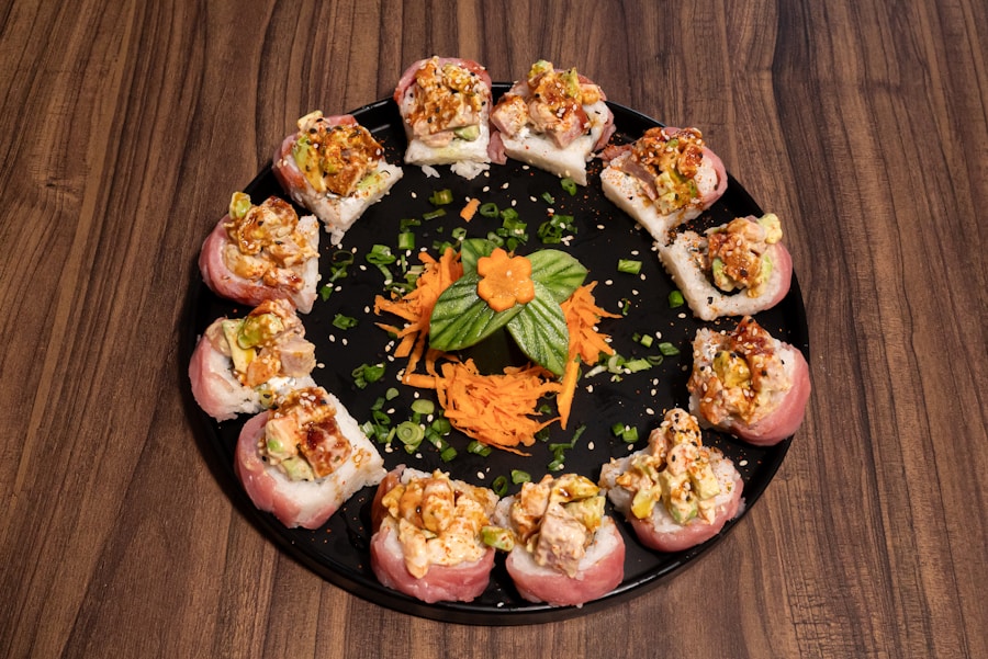 Una disposición circular de piezas de sushi cubiertas con relleno picado, alrededor de zanahorias ralladas, cebollas verdes y una flor de pepino en un plato negro sobre una mesa de madera.