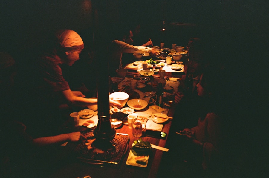 Un grupo de personas se sienta alrededor de una mesa poco iluminada, compartiendo una comida con diversos platos, bebidas y utensilios esparcidos por la mesa. Una luz cálida y tenue crea un ambiente íntimo y acogedor.