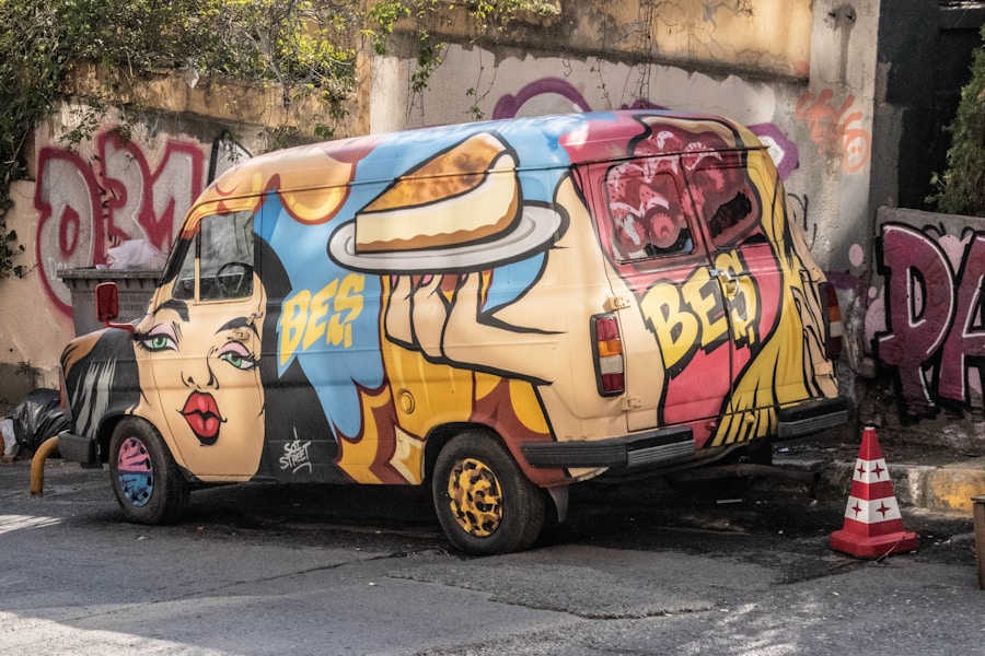 Una camioneta estacionada en la calle está cubierta de coloridos grafitis, con el rostro de una mujer de estilo pop art y una mano sosteniendo un plato de panqueques. La pared del fondo también está decorada con grafitis.