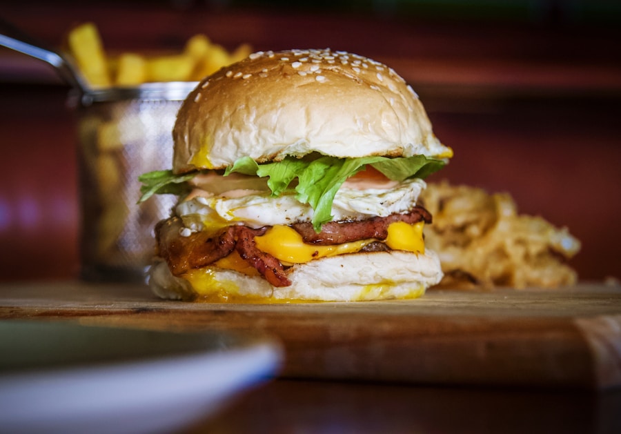 Primer plano de una hamburguesa con lechuga, tomate, bacon, queso y un huevo frito en un pan con semillas de sésamo. Al fondo, patatas fritas y aros de cebolla sobre una superficie de madera.