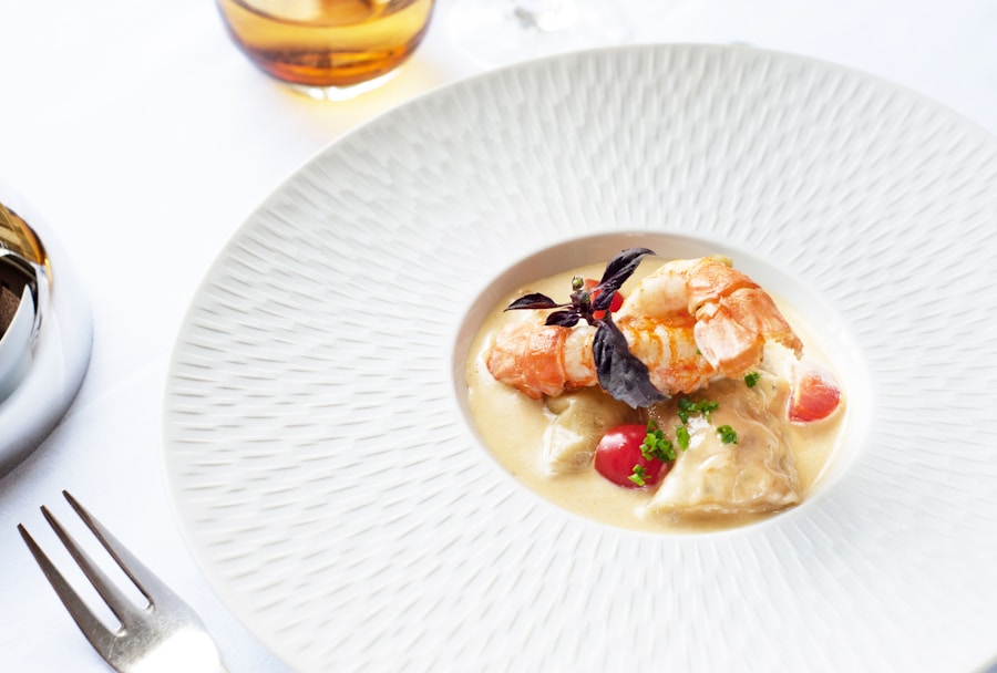 Un plato blanco con sopa cremosa de marisco, con gambas, verduras cortadas en dados, hierbas frescas y una guarnición de hojas de albahaca oscura. Cerca, sobre un mantel blanco, un tenedor y un vaso de bebida de color ámbar.