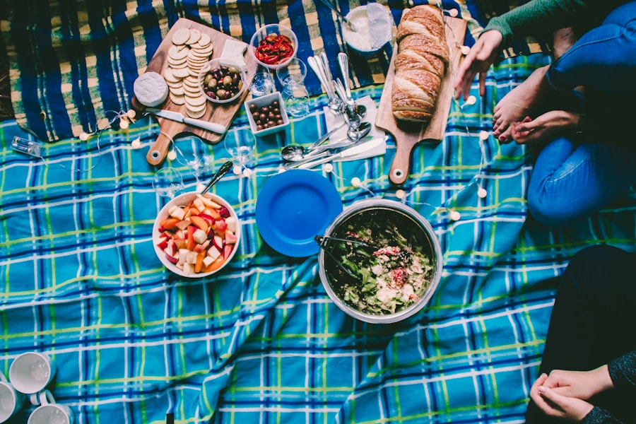Un picnic sobre una manta de cuadros azules con pan, galletas, fruta, ensalada, salsas y utensilios. Dos personas se sientan con las piernas cruzadas junto a la comida, con solo las piernas y las manos visibles.