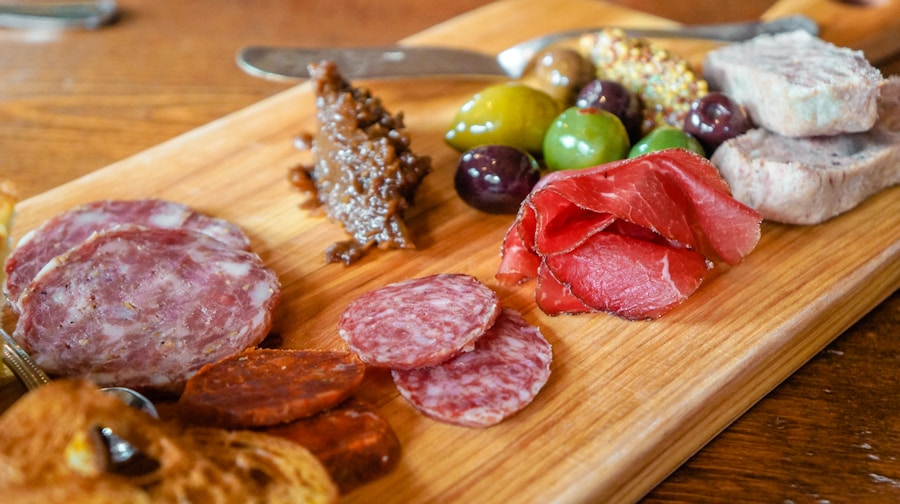 Una tabla de madera con embutidos variados, salami en lonchas, jamón serrano, paté, aceitunas, mostaza integral, chutney y trozos de pan tostado sobre una mesa de madera.