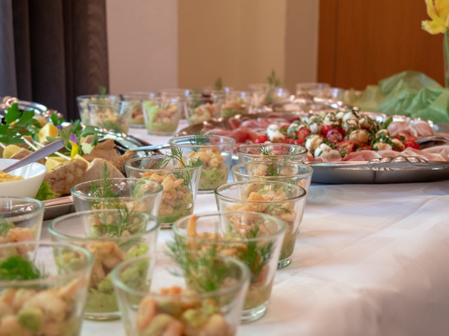Primer plano de una mesa de buffet con pequeños vasos de cristal con ensalada de gambas, adornada con eneldo, y varias bandejas de aperitivos, incluyendo brochetas de tomate y queso, dispuestas sobre un mantel blanco.