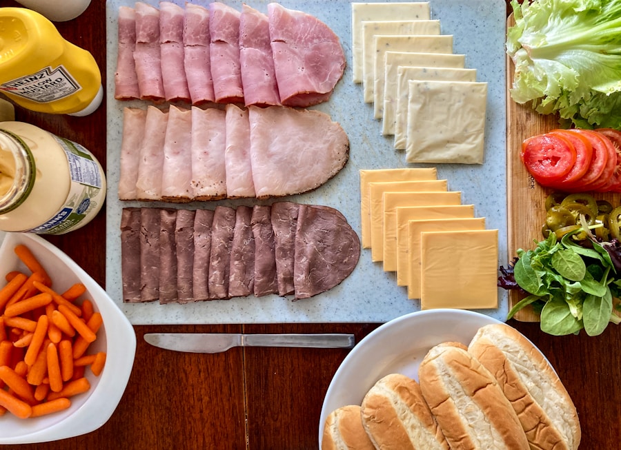 Carnes frías, lonchas de queso, lechuga, tomate, pepinillos, zanahorias baby, condimentos y panecillos para sándwich ordenados en una mesa, listos para preparar sándwiches.