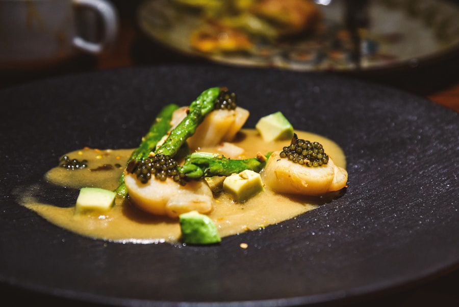 Un plato gourmet con vieiras a la plancha cubiertas de caviar, lanzas de espárragos y pequeños dados de aguacate, servido en un plato negro con una salsa cremosa.