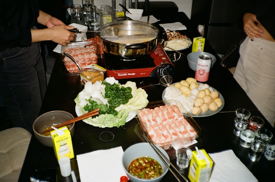 Una mesa preparada para una olla caliente con diversos ingredientes, como carne en rodajas, verduras de hoja verde, setas, albóndigas de pescado, salsas para mojar y bebidas. Dos personas preparan la comida cerca de una olla sobre un quemador portátil.