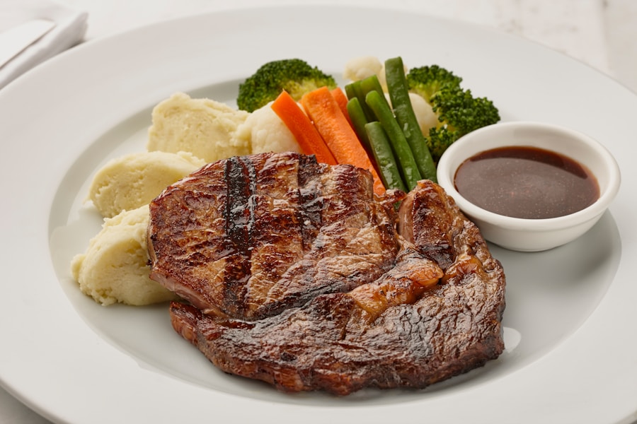 Un filete a la parrilla servido con puré de patatas, verduras variadas al vapor (brécol, zanahorias, judías verdes y coliflor) y un pequeño cuenco de salsa marrón en un plato blanco.