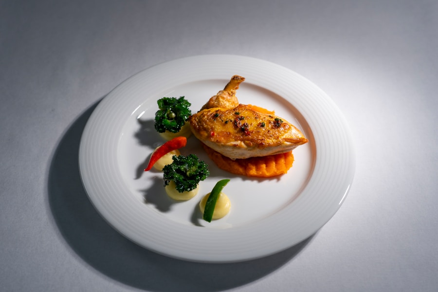 Un plato blanco con un muslo de pollo asado sobre puré de boniatos, adornado con verduras de hoja verde, rodajas de pimiento rojo y verde, y dos pequeñas porciones de salsa cremosa.
