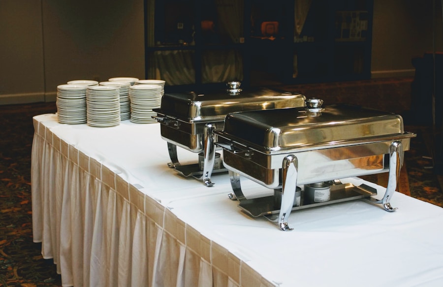 Una mesa de bufé cubierta con un mantel blanco sostiene dos fuentes metálicas cerradas y pilas de platos blancos, colocados en un banquete o evento.